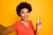 © deagreez - Self photo of cheerful charming pretty curly wavy girlfriend showing you v-sign recording video taking selfie influencing her audiences wearing orange t-shirt isolated yellow vivid color background