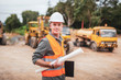 © Pituk - Caucasian handsome engineer is stand and smile holding a laptop with roll of paper on road construction vehicle background.