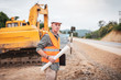 © Pituk - Caucasian handsome engineer stand holding a laptop and roll of paper on road construction site.