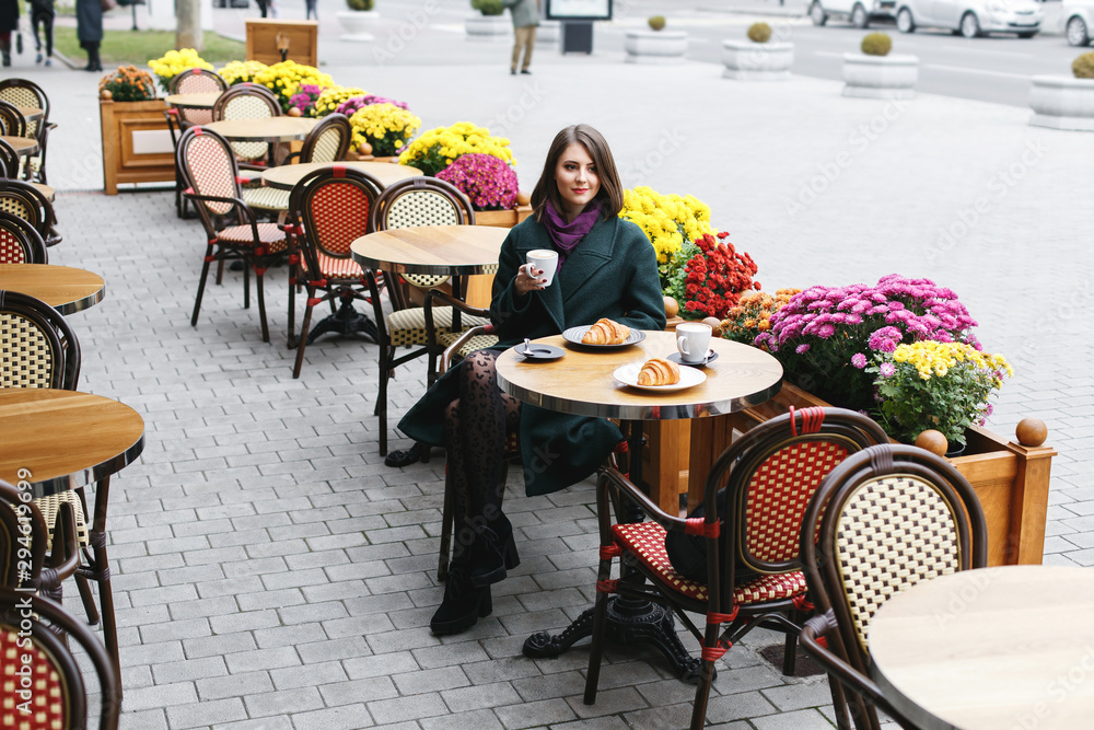 Beautiful young girl wearing green coat sitting at a table in cozy street outdoor cafe and drinking coffee with a croissant. Restaurant terrace is decorated with chrysanthemum flowers bushes in autumn - obrazy, fototapety, plakaty 