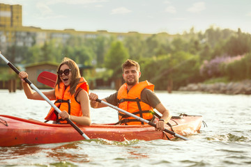 Wall Mural - Happy young caucasian couple kayaking on river with sunset in the backgrounds. Having fun in leisure activity. Happy male and female model laughting on the kayak. Sport, relations concept. Colorful.