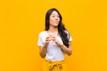 young pretty latin woman feeling proud, mischievous and arrogant while scheming an evil plan or thinking of a trick against orange wall