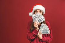 Portrait Of Beautiful Woman Wearing Santa Claus Red Costume Smiling And Holding Fan Of Money In Dollar Banknotes Isolated Over Red Background.