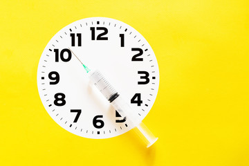 White clock dial and injection syringe indicating time against yellow background. Vaccination time. Conceptual depiction of seasonal vaccination.