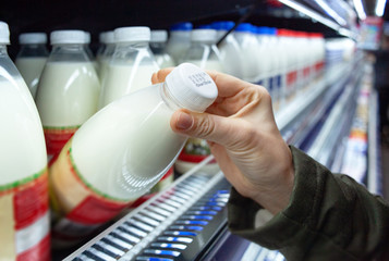 Womans hand holding milk bottle in supermarket. Man shopping milk in grocery store. Man checks product expiration date before buying it. Close-up.