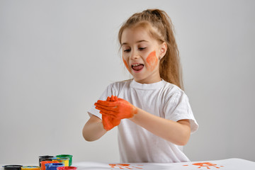 Wall Mural - Little girl in white t-shirt sitting at table with whatman and colorful paints on it, painting her hands. Isolated on white. Medium close-up.