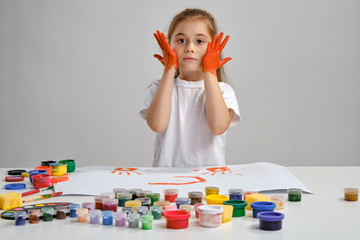 Wall Mural - Little girl in white t-shirt sitting at table with whatman and colorful paints on it, painting her cheeks. Isolated on white. Medium close-up.