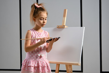 Wall Mural - Little girl artist in a pink dress is standing behind easel and painting with brush on canvas at art studio with white walls. Medium close-up shot.