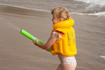 A little girl in a yellow life jacket on the beach plays with a water cannon.