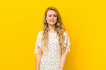 young blonde woman looking happy and friendly, smiling and winking an eye at you with a positive attitude against yellow wall