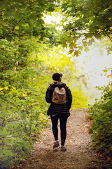Wall Mural - blond woman in dark hiking clothes and pink backpack walking in forest in autumn in park, Seen from behind, telephoto lens. Light effect on sunset. Gatineau Park, Quebec, Ontario.