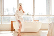 Picture of girl looking at side in white bathrobe and with towel on her head standing near bath in room with large window