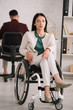 © LIGHTFIELD STUDIOS - confident, disabled businesswoman looking at camera while sitting in wheelchair in office near colleague working on background