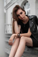 Attractive European young woman in a leather fashionable jacket in a black dress sits on the steps near a vintage white building in the city. Elegant modern girl model is relaxing outdoors. Fashion.