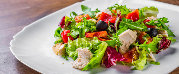 Wall Mural - Fresh salad with chicken breast, arugula, black olives,red pepper, lettuce, fresh sald leaves and tomato on a white plate on wooden table background