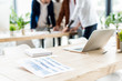 © LIGHTFIELD STUDIOS - selective focus of desk with laptop, notebook and paper with graphs and charts near businesspeople standing at workplace in office