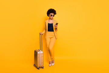 Poster - Full length photo of focused brown curly hair girl use cellphone search taxi hold bags tourism concept have abroad vacation wear style jacket pants high-heels isolated over yellow color background