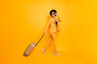 © deagreez - Full size profile side photo of cheerful positive brown curly hair girl holding trolley go walk from airport wear stylish jacket pants high-heels isolated over yellow color background