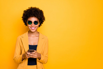 Wall Mural - Portrait of positive cheerful afro american girl in sunglass use her smartphone type text post comments feedback read feednews wear stylish trendy suit clothes isolated over yellow collor background