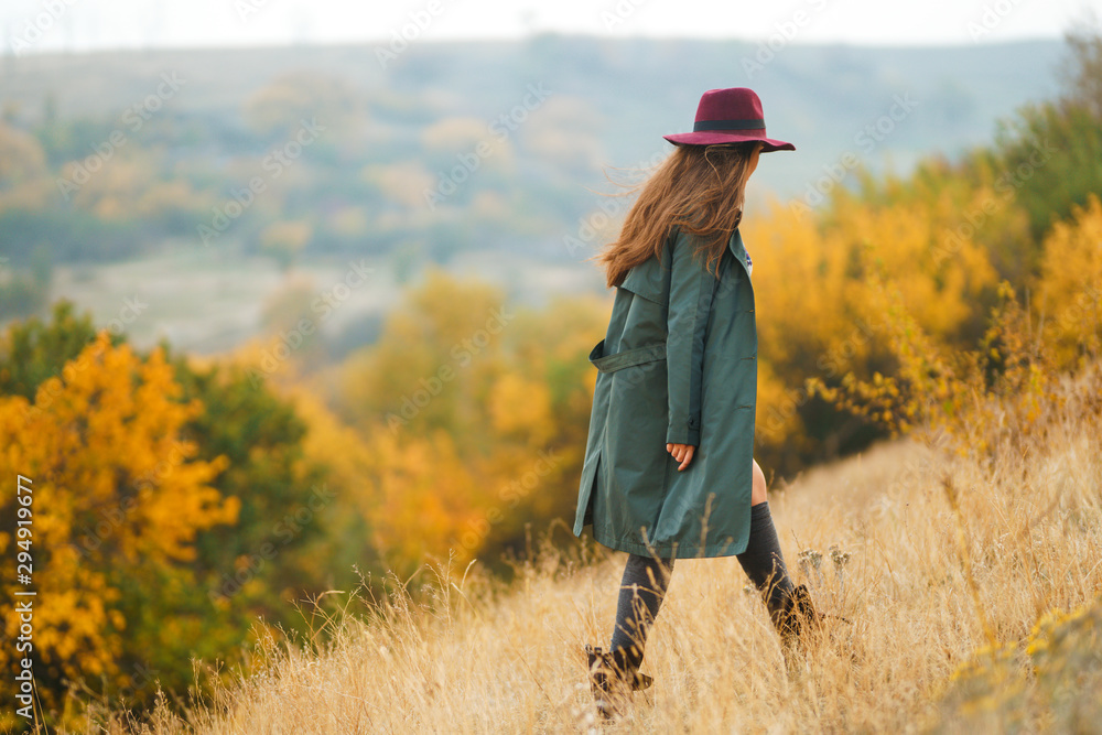Beautiful young stylish girl in a coat walks in the autumn in the park. The girl is dressed in a green coat and a red hat. Beautiful evening. Autumn fashion. Lifestyle. High fashion portrait.  - obrazy, fototapety, plakaty 