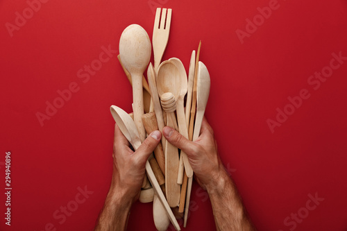 Fotomural cropped view of man holding wooden spoons, fork and kitchenware on red backgroun