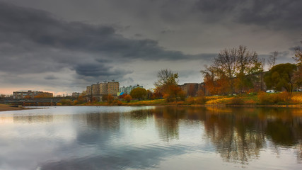 Autumn city landscape trees with colorful foliage are reflected in the river against a cloudy sky lit by the rays of sunset.