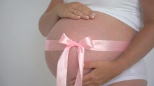 Close Up Of Woman Pregnant Belly With Pink Ribbon Bow. Mom Anticipation Expecting Baby. Pregnant Woman Touching Belly On White Background. Newborn Girl. Pregnancy Maternity Motherhood Mother Hands 4 K
