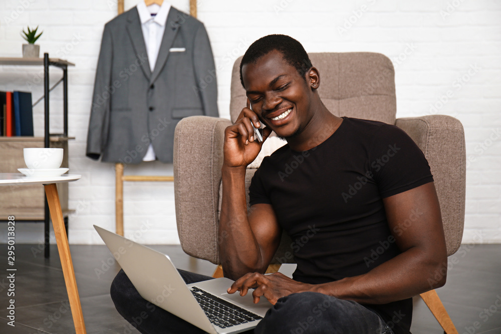 Handsome African-American man with laptop talking by phone at home