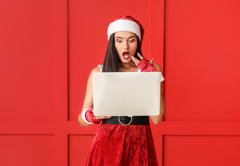 Wall Mural - Surprised young woman in Santa Claus costume and with laptop on color background