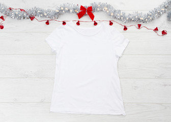 Close up white blank template t shirt with copy space and Christmas Holiday concept. Top view mockup t-shirt and red bow on white wooden background. Happy New Year decorations accessories. Xmas outfit