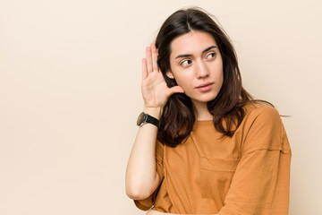 Wall Mural - Young brunette woman against a beige background trying to listening a gossip.