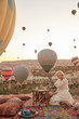 © monchak - a girl in white dress sitting on old turkish carpet with pillows and looking on many air balloons in the sky in Cappadocia
