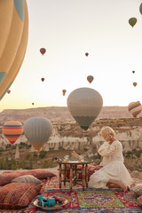 a girl in white dress sitting on old turkish carpet with pillows and looking on many air balloons in the sky in Cappadocia