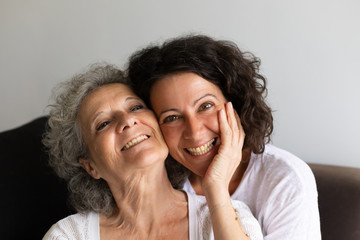 Cheerful senior mother and adult daughter posing at home. Happy elderly lady touching cheek of middle aged woman, laughing and smiling at camera. Affection concept