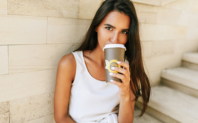 Canvas Print - Beautiful brunette woman holds disposable cup of coffee in the city street. Pretty female takeaway coffee in hand. Pretty student drinking coffee outdoors in the morning.