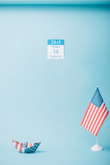 Sticker - paper calendar with Columbus Day inscription near paper boat and American national flag on blue background