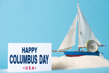 Sticker - card with happy Columbus Day inscription near miniature ship in white sand isolated on blue