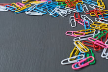 Lot Of Scattered Different Colors Paper Clips Red, Green, Blue, Yellow, White And Pink For Office Work Or Education Lies In Corner On Dark Scratched Concrete Table. Space For Text