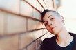 Young and attractive girl with shaved head and piercings. He has his head resting on a brown brick wall and looks at the camera with a nostalgic face. It is an urban scene.