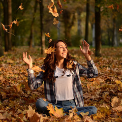 Beautiful girl sitting on the ground and throwing yellow leaves in park. Leisure time on warm autumn day. Autumn mood. Enjoy season