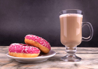 A composition of two pink glazed donuts and a glass cup of coffee with milk