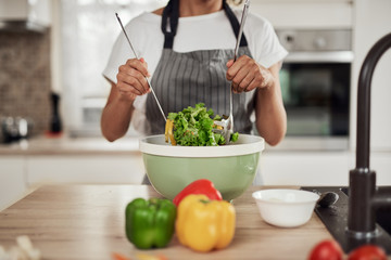 Wall Mural - Attractive mixed race woman in apron mixing vegetables in bowl while standing in kitchen at home.