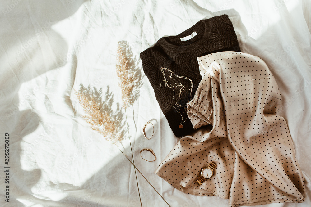Woman trendy fashion clothes on white linen bed. Flat lay, top view. Black sweater, silk skirt with polka dots, gold necklace, watch, earrings, reeds. Feminine french style concept. - obrazy, fototapety, plakaty 