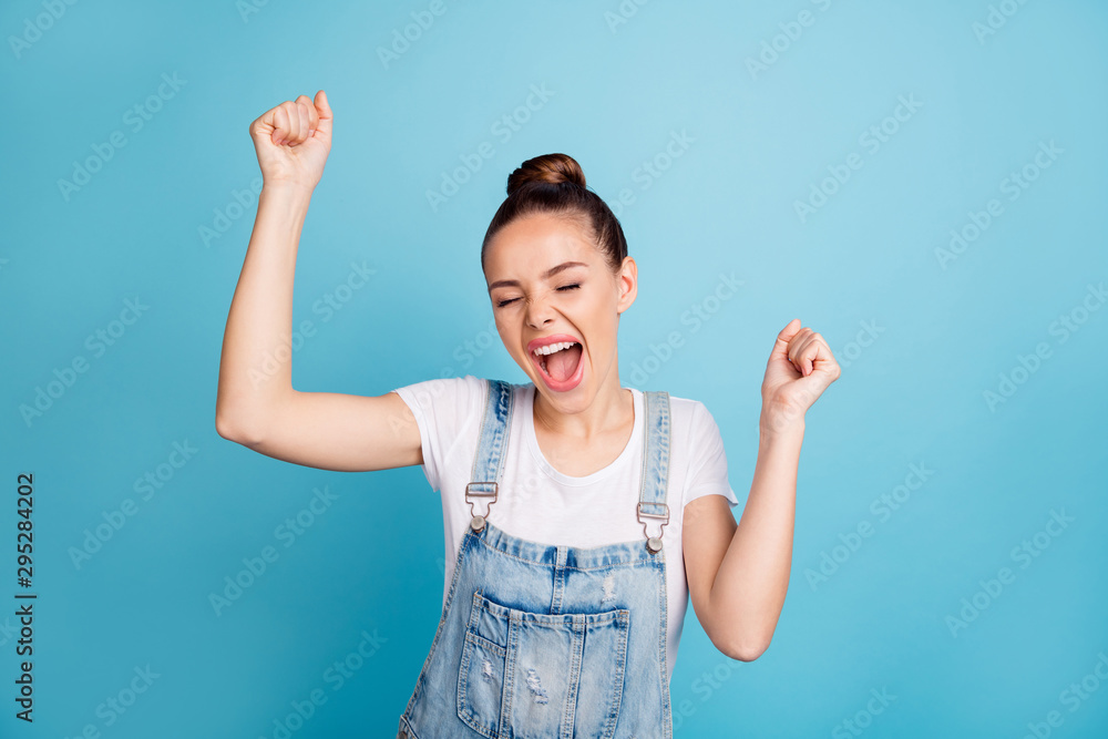 Portrait of cheerful lady with her eyes closed raising fists yelling yeah wearing white t-shirt denim jeans overalls isolated over blue background - obrazy, fototapety, plakaty 