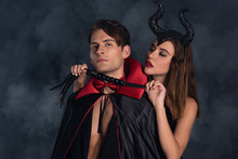 Attractive Woman With Horns Holding Flogging Whip Near Man In Vampire Halloween Costume On Black With Smoke