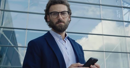 Young confident Professor in Glasses looking to the Camera confidently. Portrait of young Caucasian Man with stylish hair and Look. Using his Smartphone. Social Network. Work outside. Success. People.