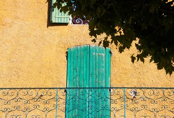 View beyond tree on stone facade of typical French house and balcony with ancient ornate lattice work and green door shutter - Castellane, France