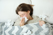 © Photoboyko - A young woman lying in bed under a blanket and blowing her nose in a napkin.Seasonal cold or flu concept
