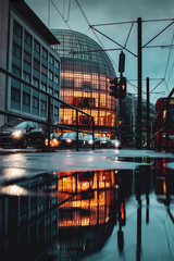 Wall Mural - Rainy moody urban street view in down town on a cold and dark day. Modern illuminated architecture building with water reflection in the puddle with tram train. Cologne, Germany