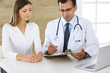 © Iryna - Doctor and patient discussing the results of a physical examination while sitting at a desk in a clinic. A male doctor using a clipboard to fill out a medical history of a young woman's medication